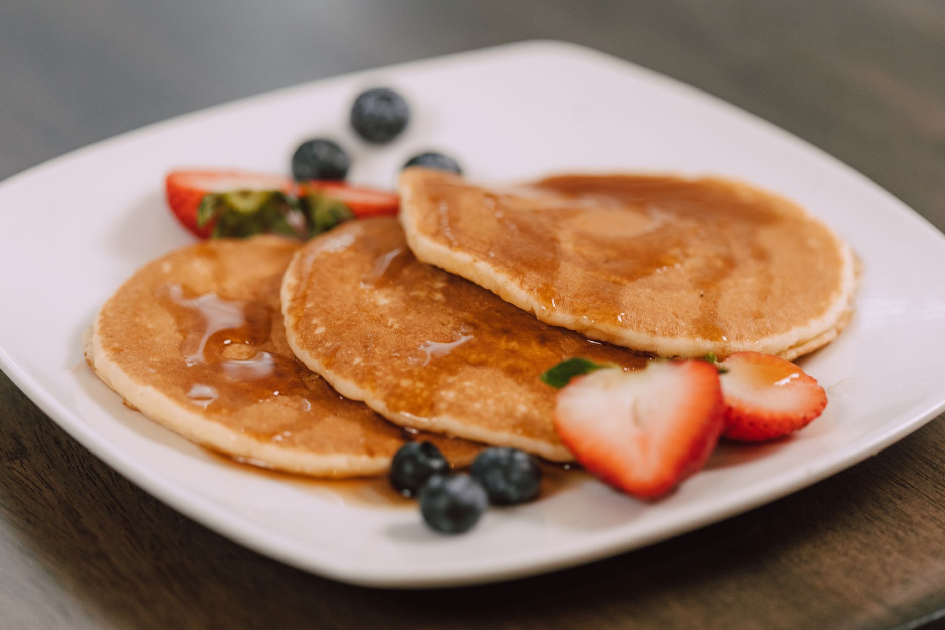 Assiette blanche de trois pancakes nappés de sirop, avec des fraises coupées et des myrtilles, sur une table en bois sombre.