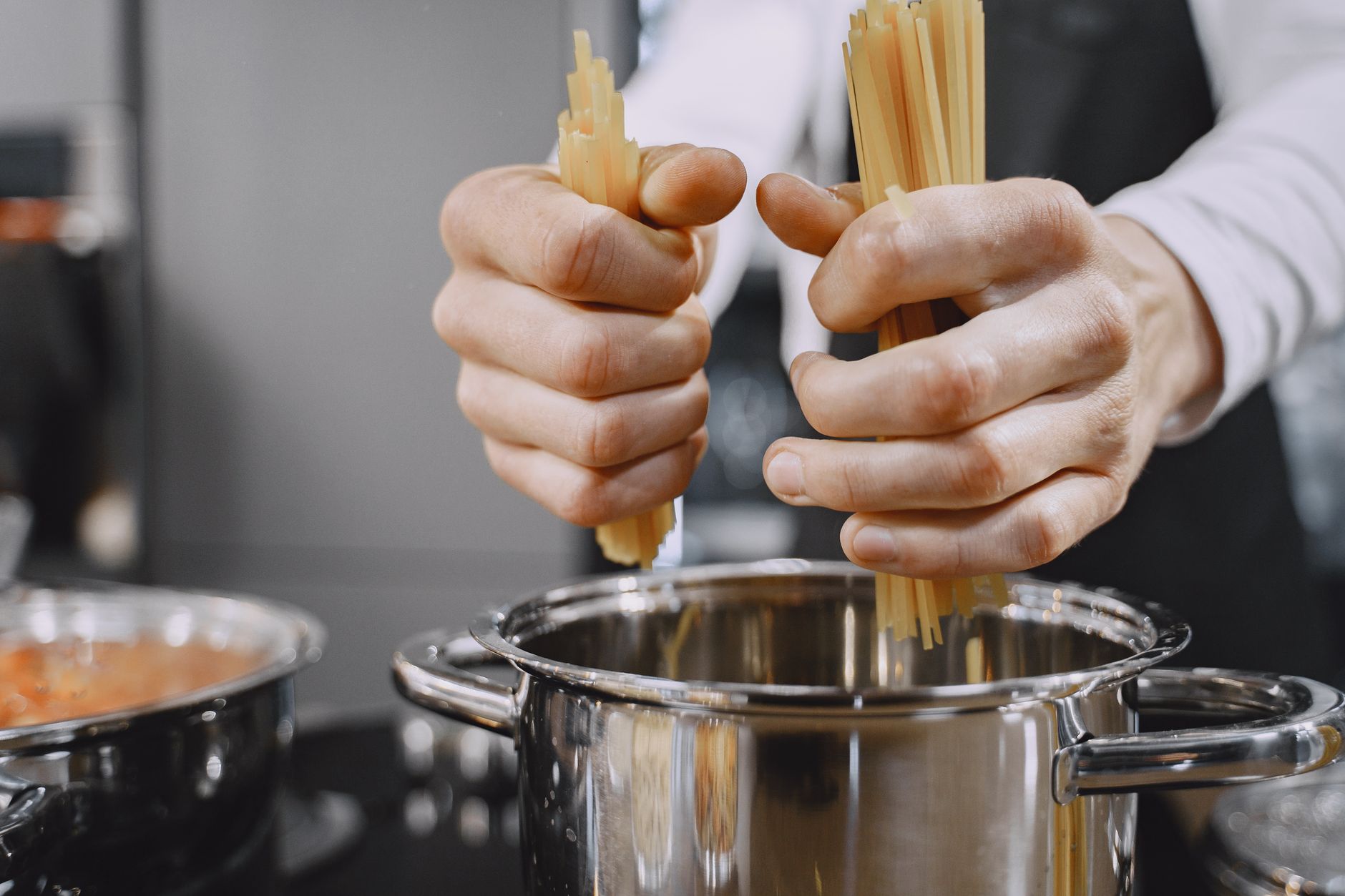 Deux mains plongent des pâtes sèches dans une casserole en inox, dans une cuisine, avec un second récipient au premier plan à gauche.