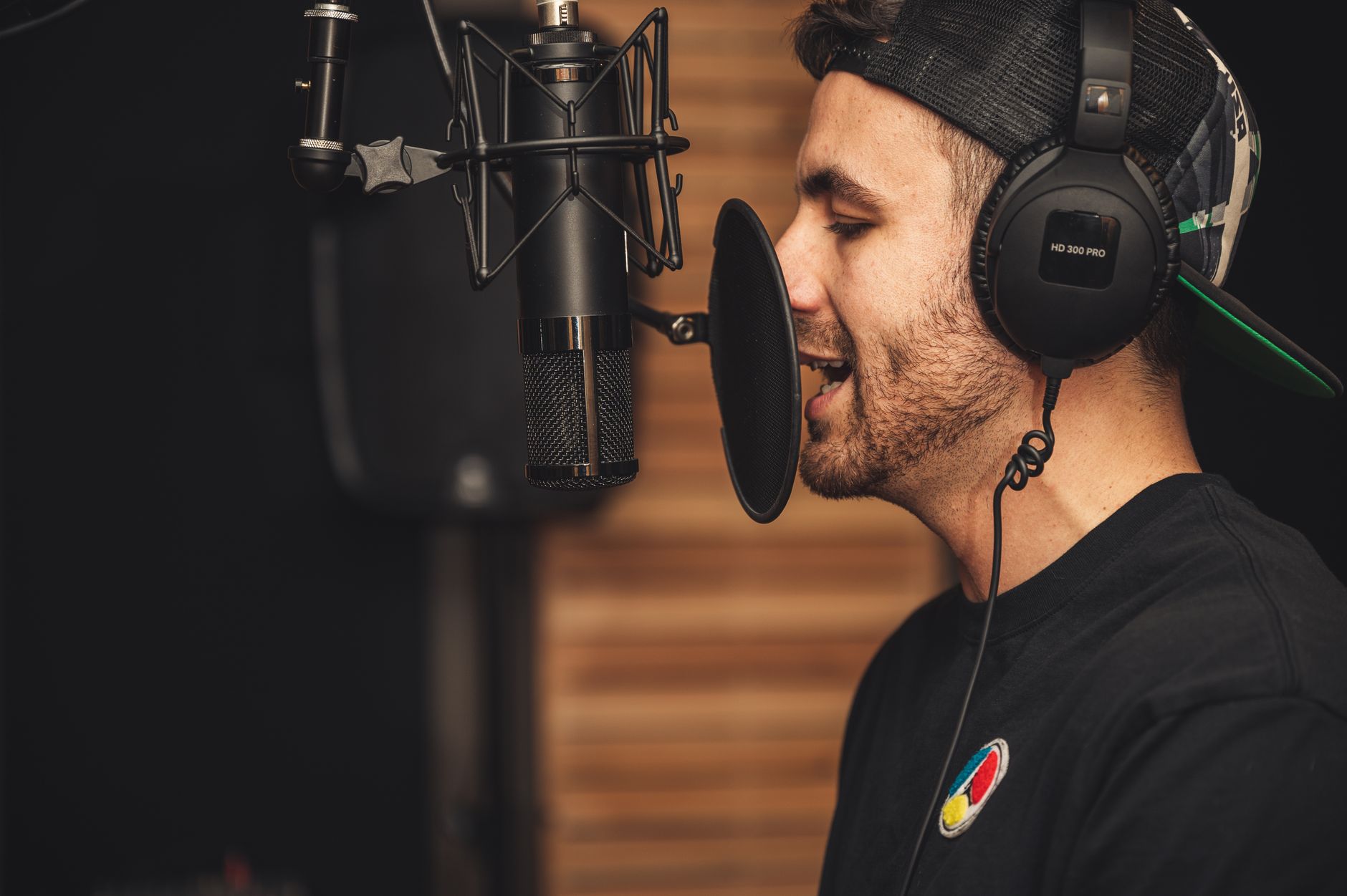 Homme en casquette et casque audio parlant devant un microphone de studio avec filtre anti-pop, devant un mur en bois.
